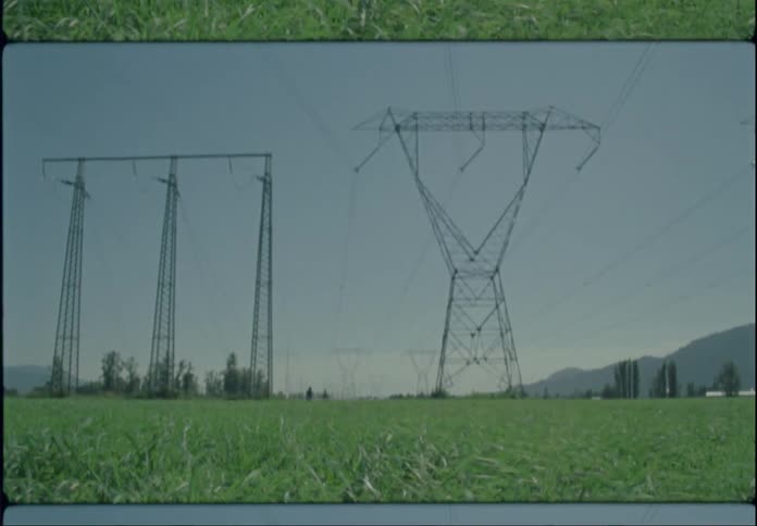 Two people are walking near utility towers as the grass moves in the breeze and a mountain in the distance