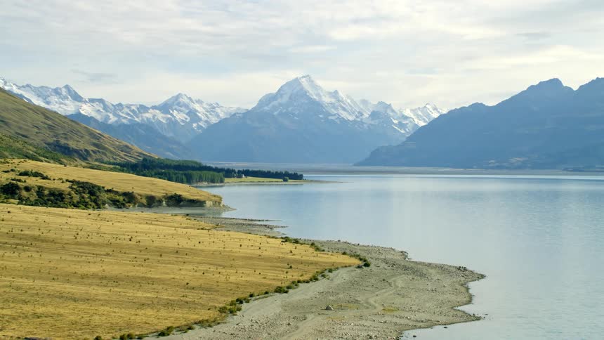 Scenic native grasses grow at the foot of a mountain along the lake edge with snow covered mountains in the distance under a cloudy sky