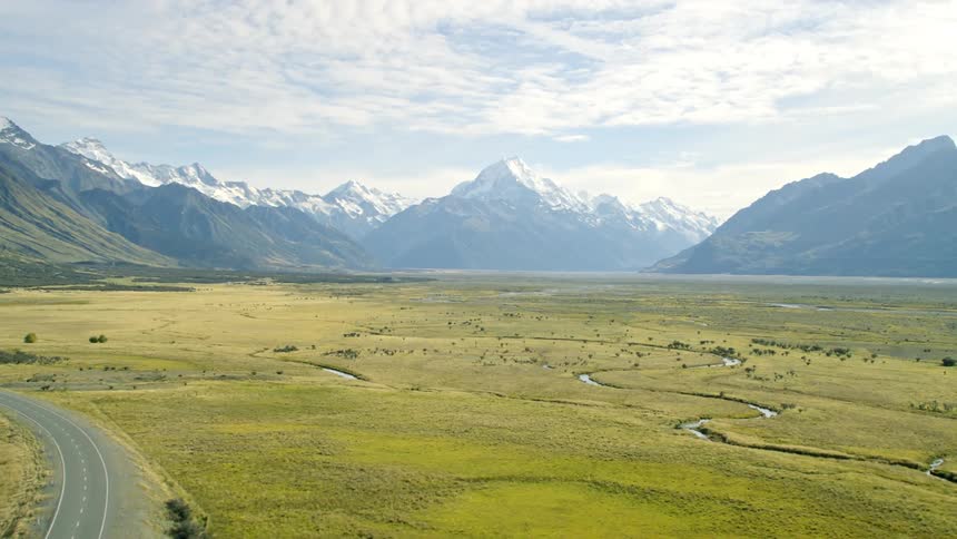 A scenic road in a vast grassland and in the background huge mountains with New Zealand famous mountain