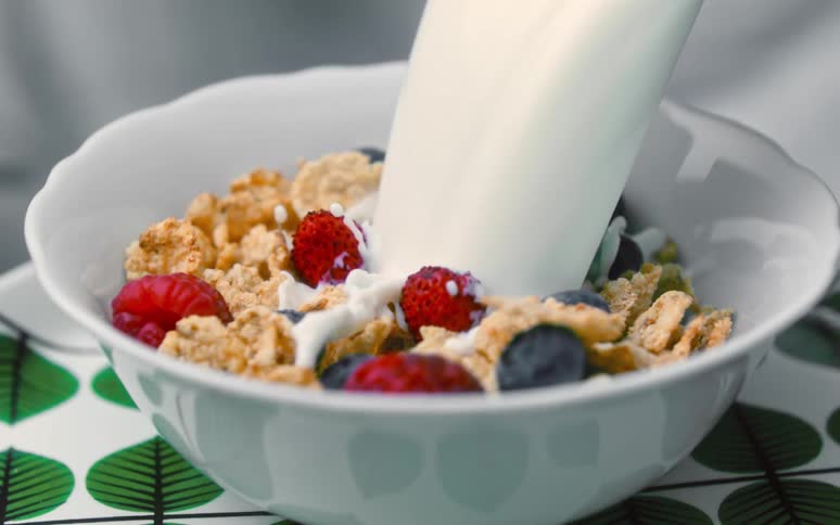A bowl of cereal with blueberries and strawberries is on a table as milk splashes as it pours into the bowl