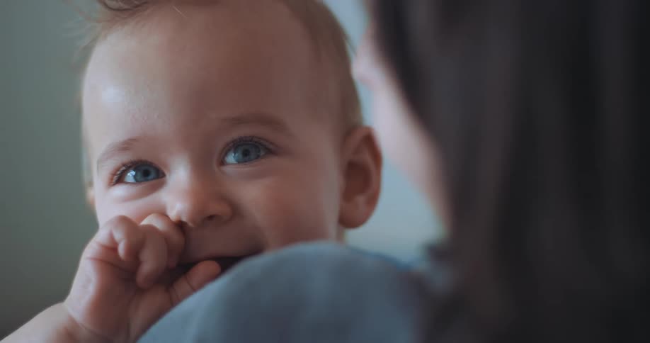 Baby mouths hand and smiles while looking over mothers shoulder