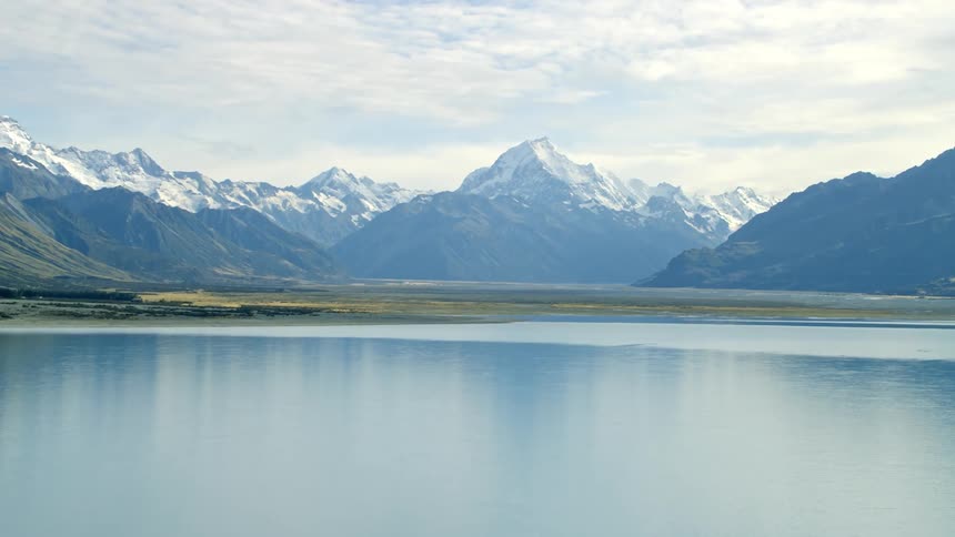 The sun shines upon icy water along the lake with snow covered mountains in the distance under a cloudy sky