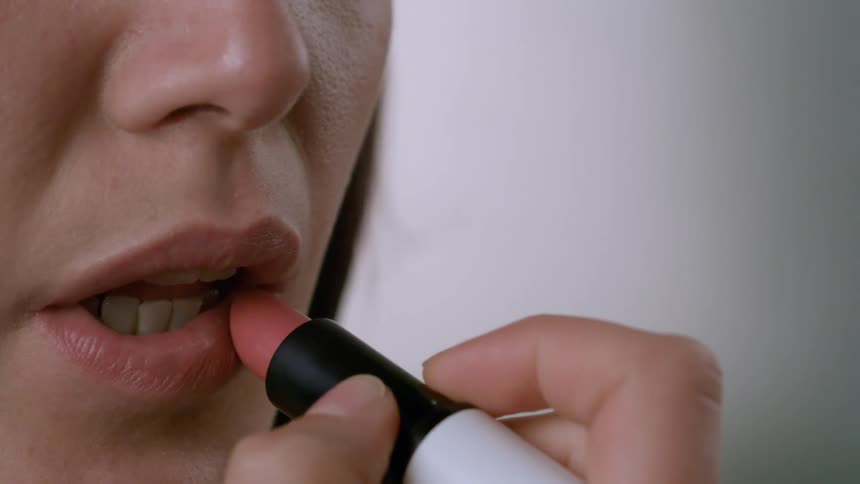 A woman holding a tube of lipstick slowly applies it to her lips