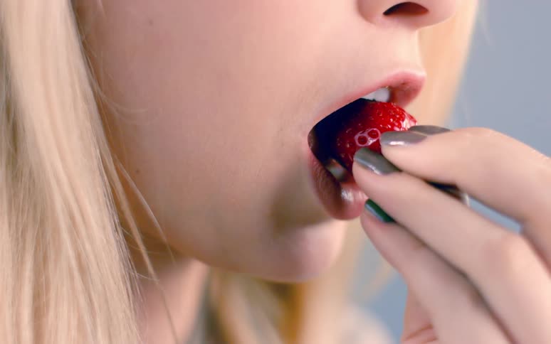 A girl holds a strawberry and places it in her mouth as she takes a bite