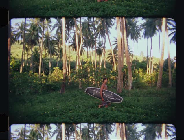 A shirtless boy carrying a board in his arms walking through tropical vegetation