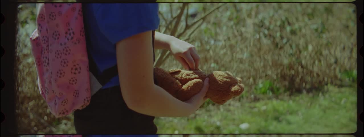 A little girl with a pink backpack picks up a teddy bear from the ground
