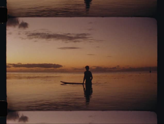 Man with his back to the camera in the sea with his surfboard under a reddish sky