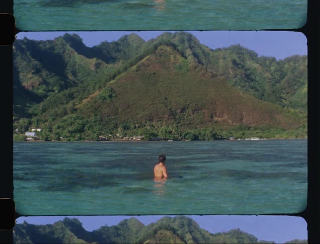 A man walking through crystal clear water next to a green mountain