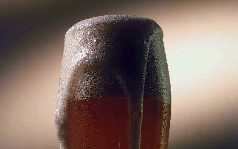 Beer suds overflow on the sides of a tall glass in slow motion