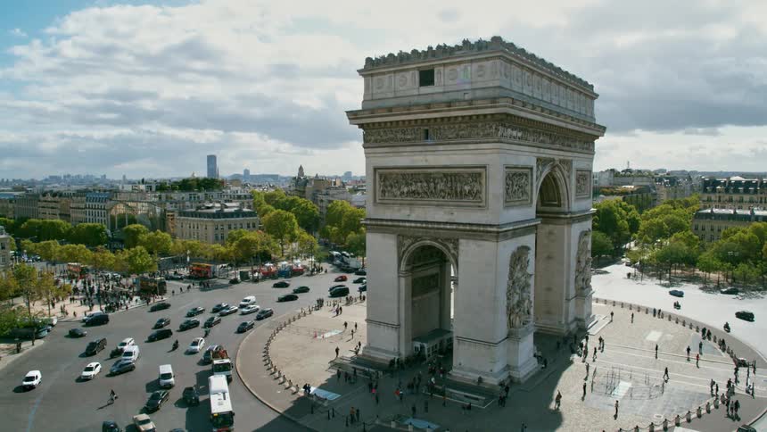 Pedestrians visit arc de triomphe as traffic flow along roundabout encircling it on a cloudy day