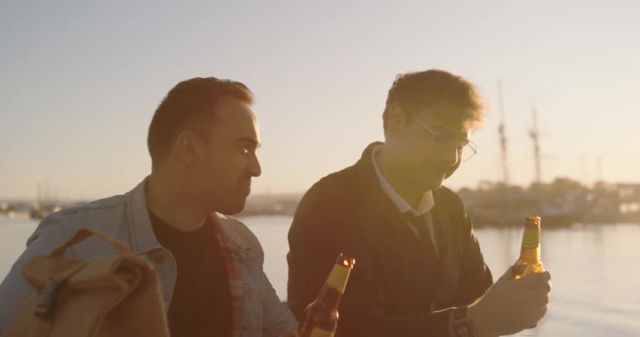 Two men are sitting at a picnic table along the coast drinking beer as the sun shines brightly over the water