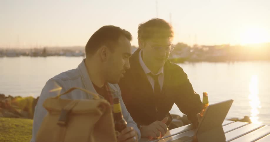Two men hold discussion over laptop while sitting at picnic table on shore with beer bottles