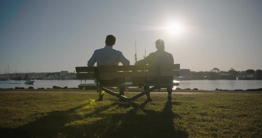 Two men are sitting on a bench along the coast overlooking the water and boats as two people walk past them