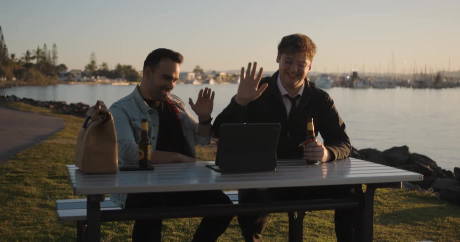 Two men sitting outdoors during video conference wave at laptop screen