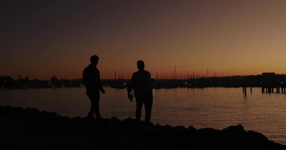Two friends throwing pebbles in the bay while enjoying a beer on the shore at dusk