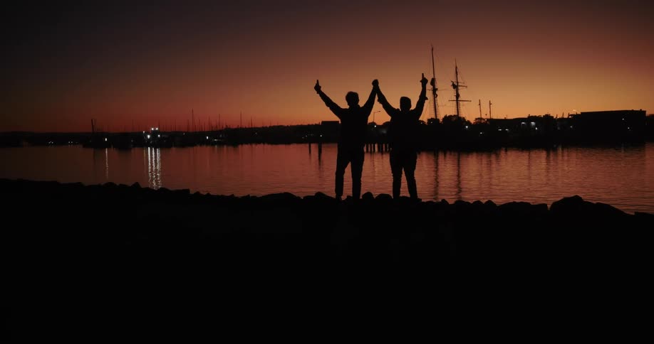 The silhouettes of two men cheer at a lake at sunset