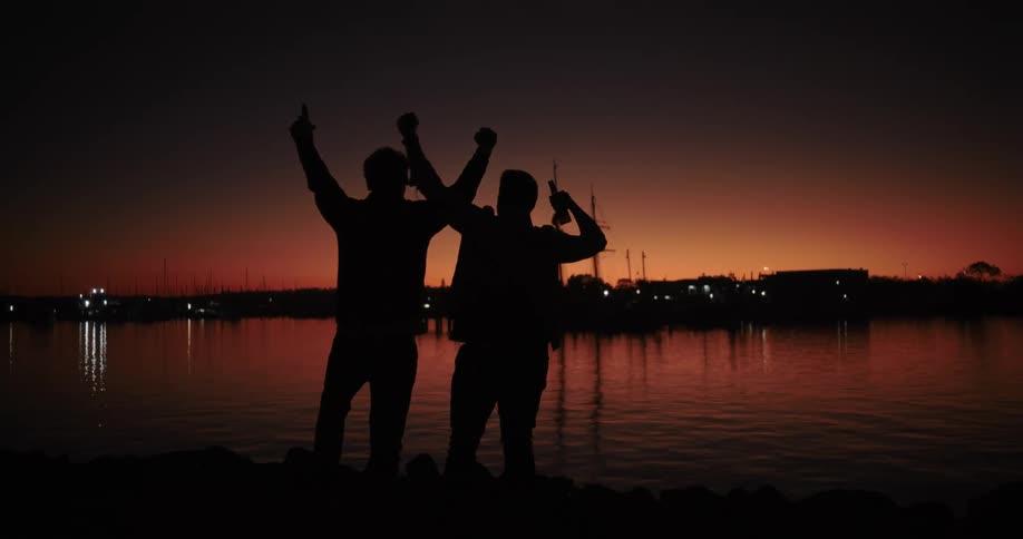 Two men celebrate on lake shore while silhouetted against afterglow at night