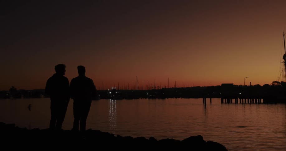 Two men facing a lake on an orange morning and one of them throws something into the water