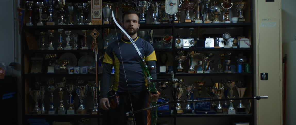 A man stands in front of a trophy case with a bow and arrow in hand