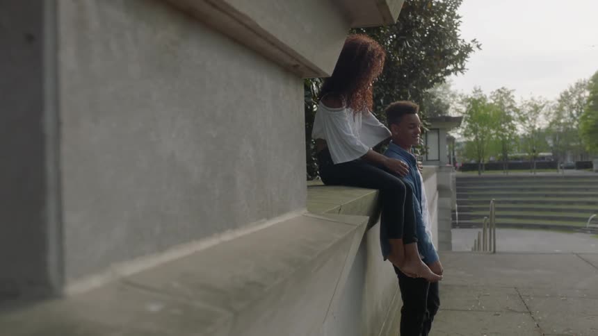 A woman sits on a wall with her hands on a man's shoulders
