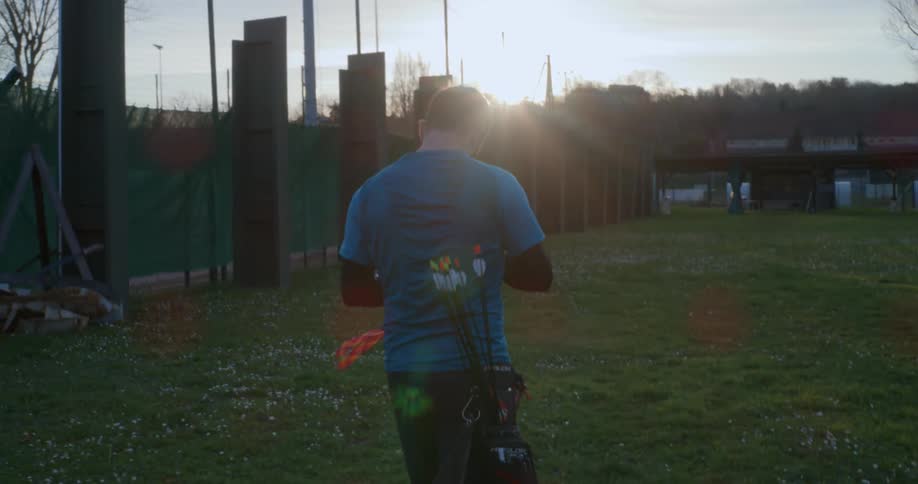 A young man carrying a bag of arrows walks across a sports field at sunset