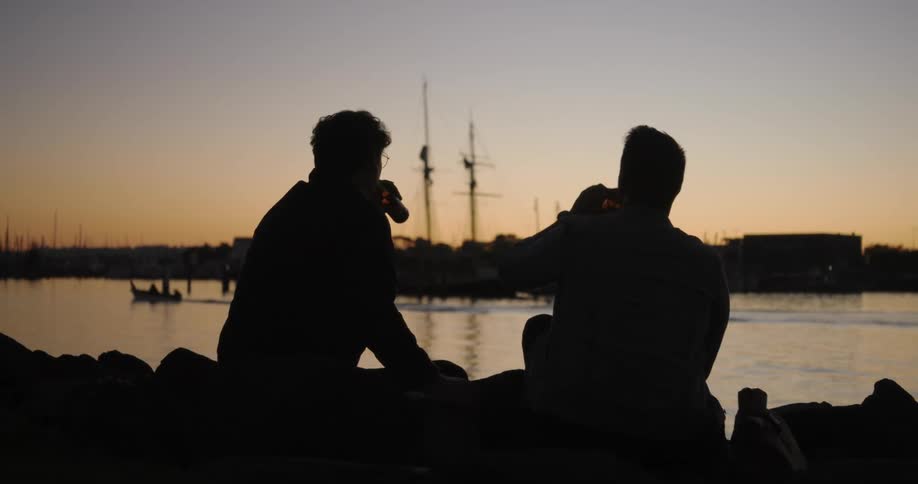 Two friends enjoying beer while sitting by the waterfront at sunset