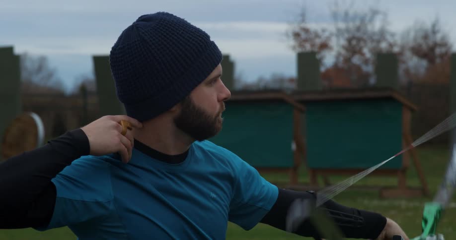 A bearded man wearing a beanie practices his archery skills on a cloudy fall day