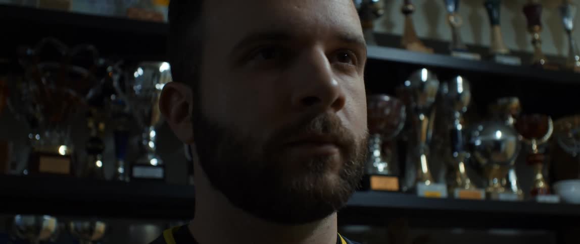 A bearded man staring straight ahead behind him is a wall of trophies