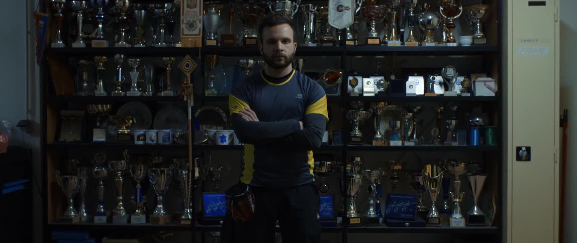 A bearded man in black and yellow clothing with his back to a wall of trophies and staring straight ahead