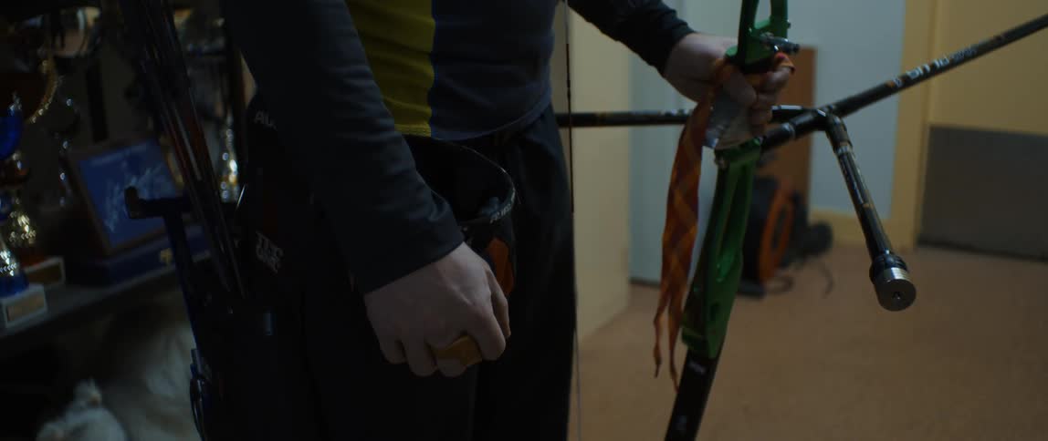 A man standing in the doorway of a dark room is holding a bow in his hand as he looks into the hallway