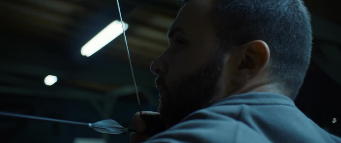 A bearded man wearing a gray jacket shoots a bow and arrow in a garage