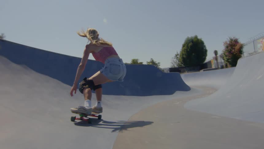 Long haired blonde woman wearing denim shorts is riding a skateboard on a course during a sunny day