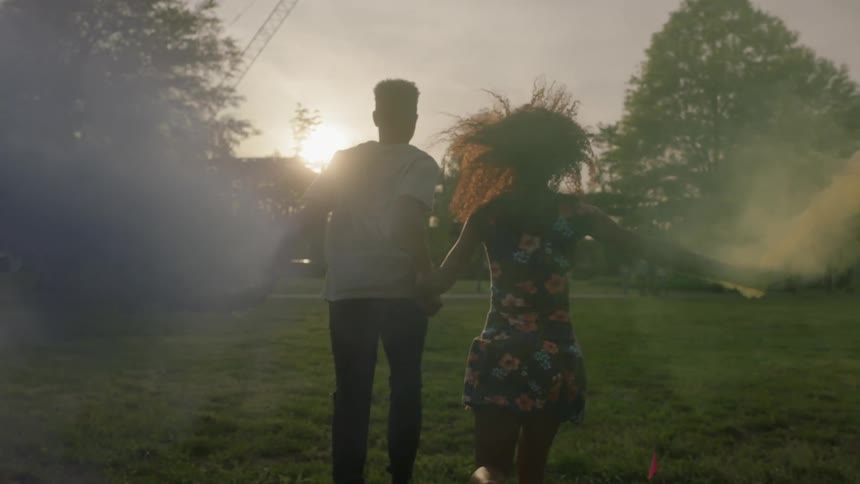 Two people run over a grass field with smoke cans