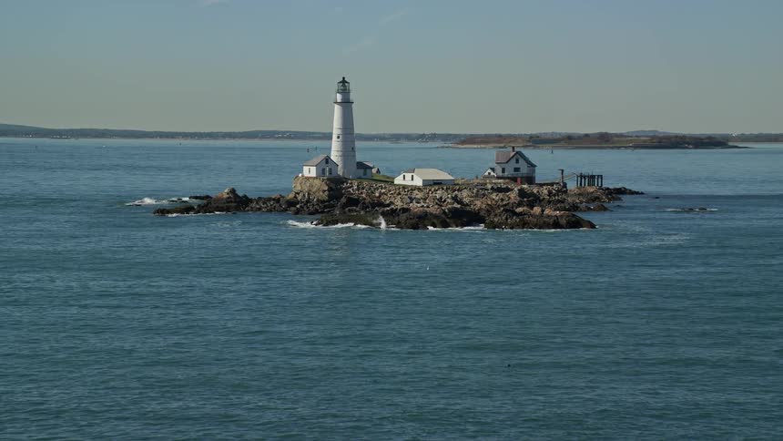 Waves splash against rocks on little brewster island with the boston lighthouse and a few houses