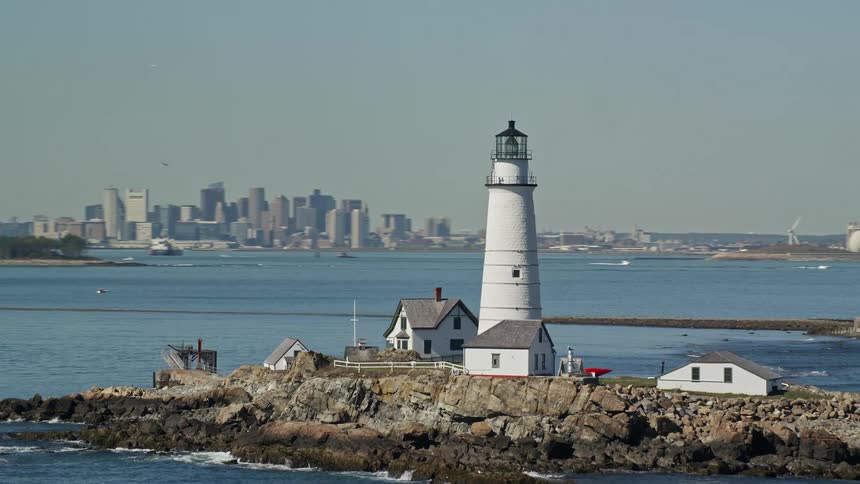 The boston lighthouse stands tall on little brewster island with several houses overlooking the atlantic ocean and the city skyline in the distance