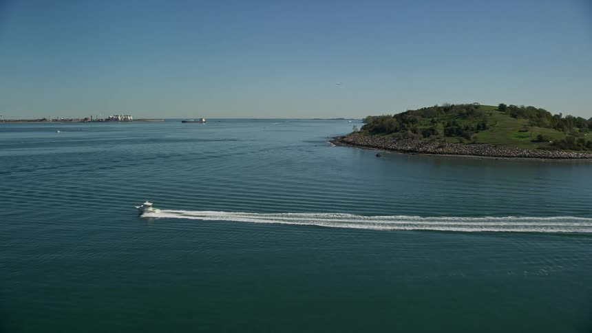 A small boat sails across the water near the coast as and airplane flies over the lake and a carrier ship is in the distance