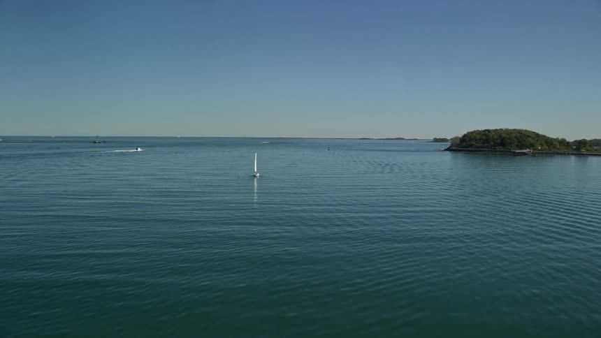 A small boat sails across the water near the coast among a sailboat and a carrier ship