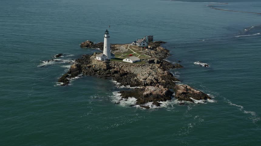 Waves splash against rocks on little brewster island with the boston lighthouse and houses