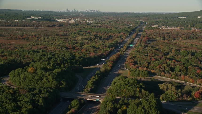 Traffic moves on highways outside of neighborhoods and woods with trees in fall colors with tall buildings and skyscrapers on the city skyline in the distance