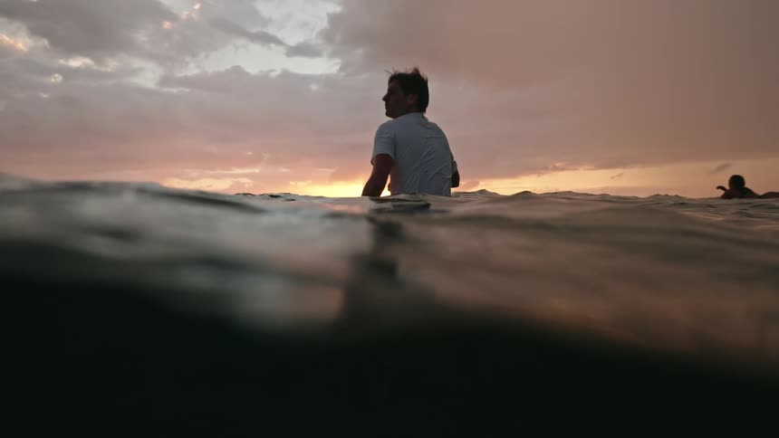 A man sitting on a surfboard in the middle of the sea observing nature with a reddish sky