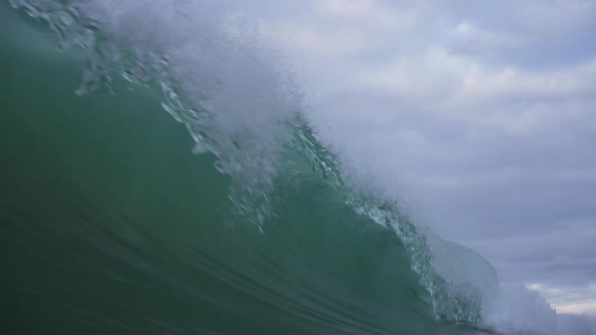 A huge waves splashes as it curls and rolls into shore under a cloudy sky and the view moves underwater