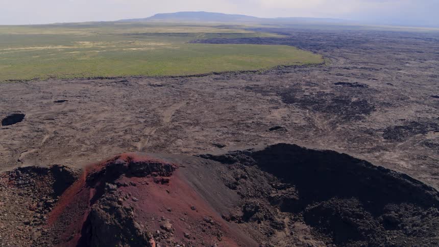 A large crater of red volcanic rock sits along the edge of a volcanic lava field that extends into sagebrush steppe rangeland