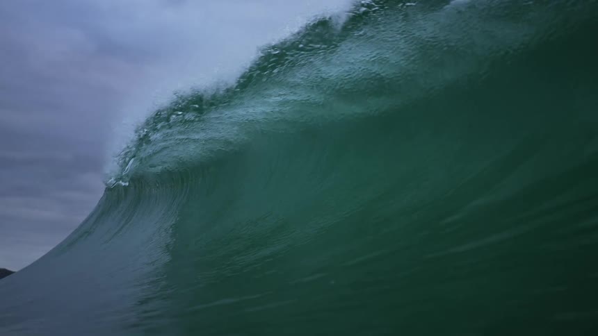 A wave is rolling into shore under a cloudy sky as it splashes and curls and rolls overhead