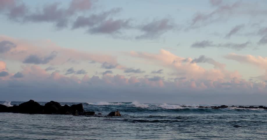 Waves splash against rocks along the coast as they roll into shore under a cloudy sky