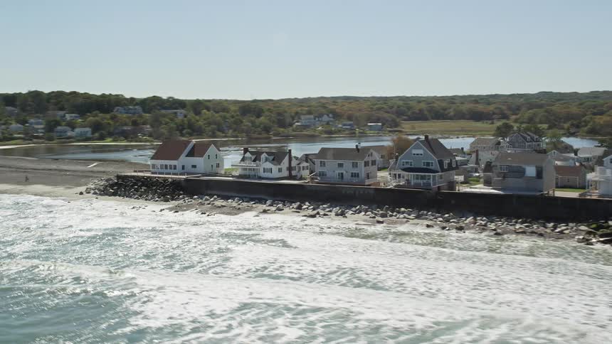 The sun shines upon coastal homes as waves roll into shore with suburban neighborhoods in the distance