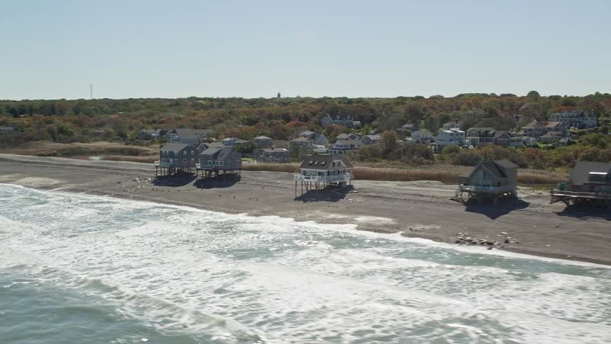 Houses stand on stilts across the beach as waves roll into shore on the outskirts of suburbs with houses and trees in the distance