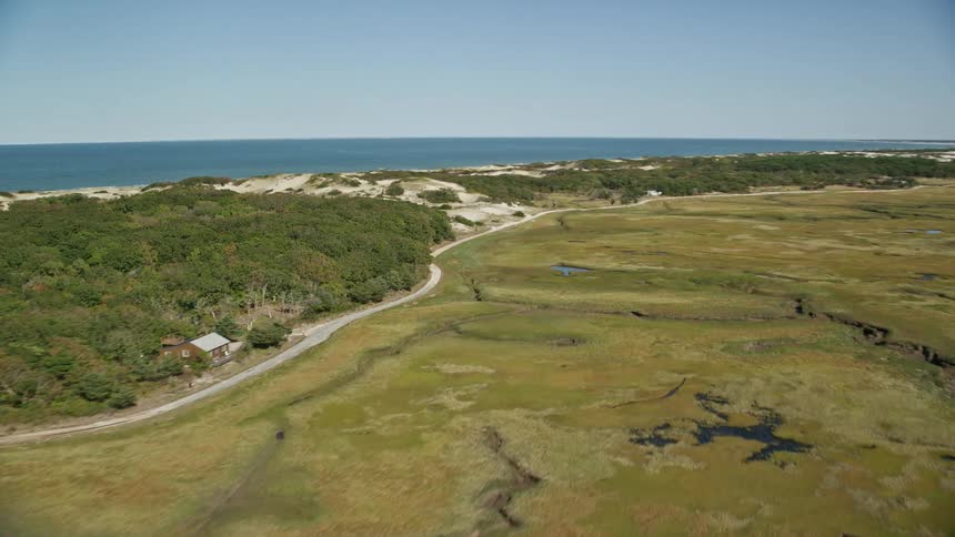 A long road runs near hills and woods along the coast as the ocean stretches out to the horizon