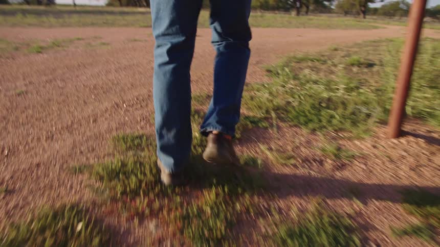 The legs of a person wearing blue jeans and brown boots are seen walking alongside a dirt road with houses in the distance