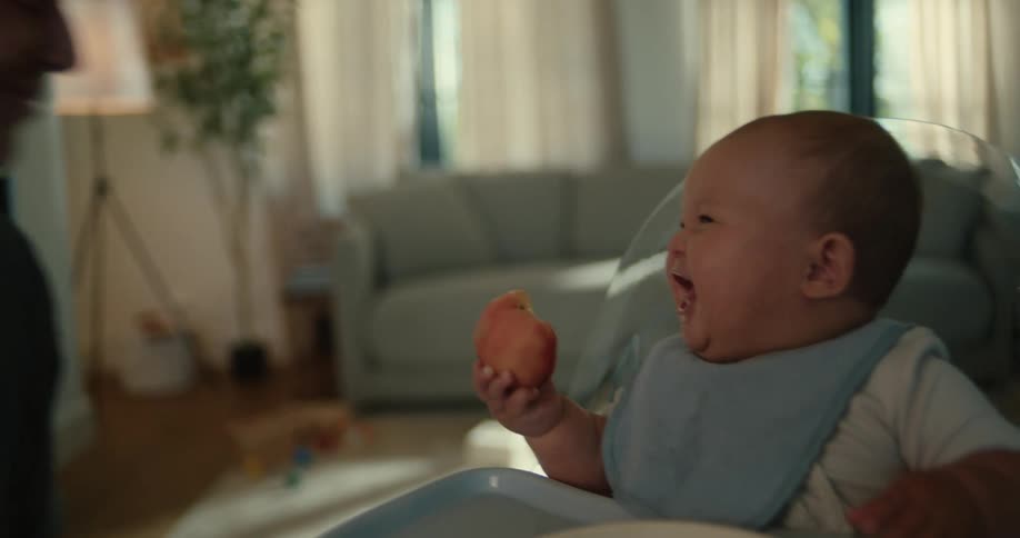 Baby sitting down next to a table eating a pastry and laughing and having fun as a thier parent pretends to bite the food from their hand