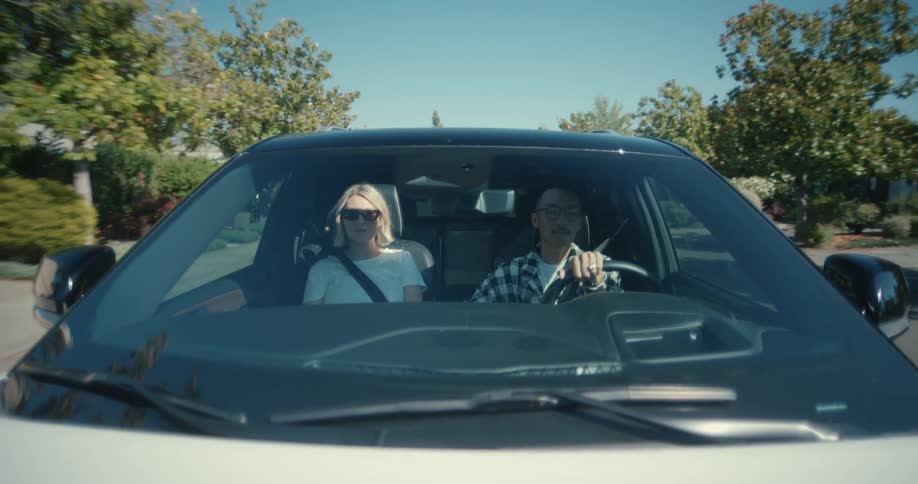 A man and a woman driving in a car through the street filled with trees nearby houses